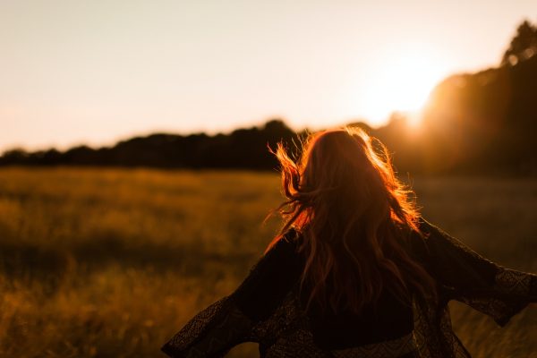 Person with long hair runs through a field at sunset, with the sun casting a warm glow.