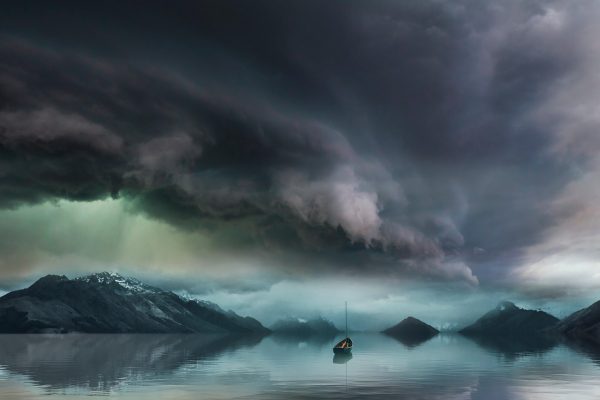 A small boat on calm water beneath dramatic, dark storm clouds, surrounded by mountains.