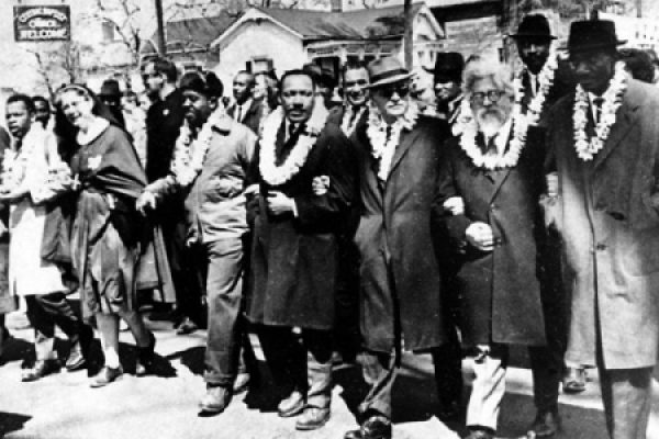 A group of people link arms while marching in a civil rights protest, wearing leis and coats, with buildings in the background.