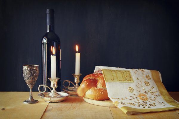 Wine bottle, goblet, lit candles, and challah bread covered with a decorative cloth on a wooden table.