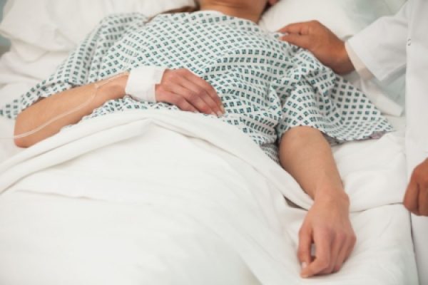 Patient lying in hospital bed with an IV drip, doctors hand resting on their shoulder.
