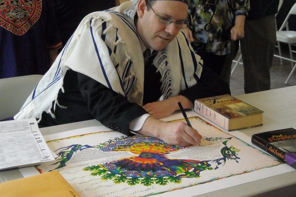 A man in a prayer shawl signs a colorful document at a table with books and papers.