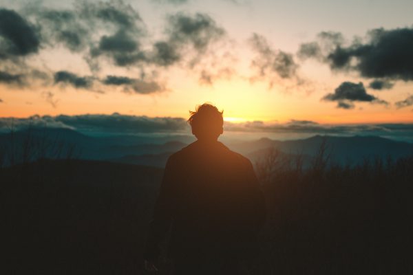 Silhouette of a person standing against a sunset over mountains, with clouds in the sky.