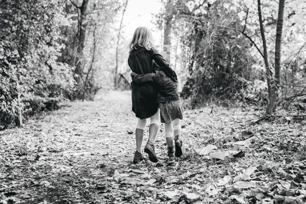 Two children walk arm in arm down a leaf-strewn forest path in black and white.