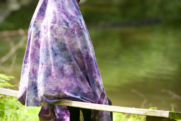 A person in a purple floral shawl stands on grass by a wooden bench near a pond.