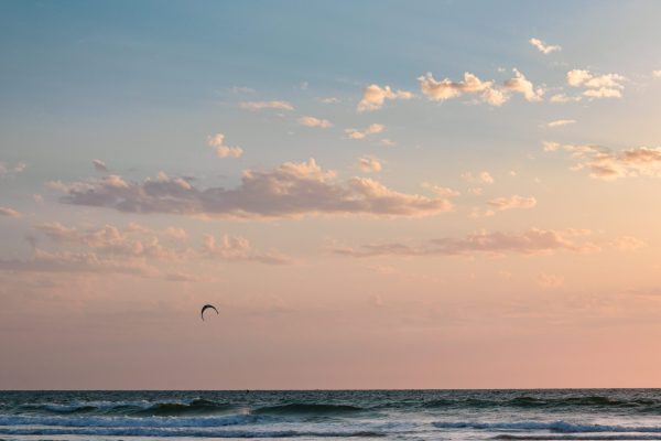 A calm ocean at sunset with gentle waves, a kite surfer in the distance, and clouds in the sky.