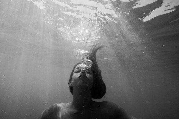 Black and white image of a person underwater, bubbles rising from their mouth, and light beams filtering through.