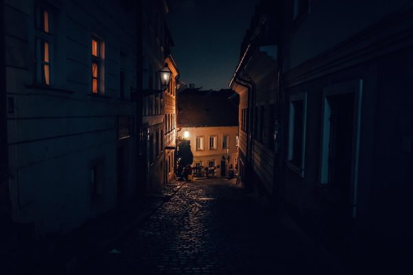 Dimly lit narrow street at night with cobblestone path, old buildings, and a glowing streetlamp.