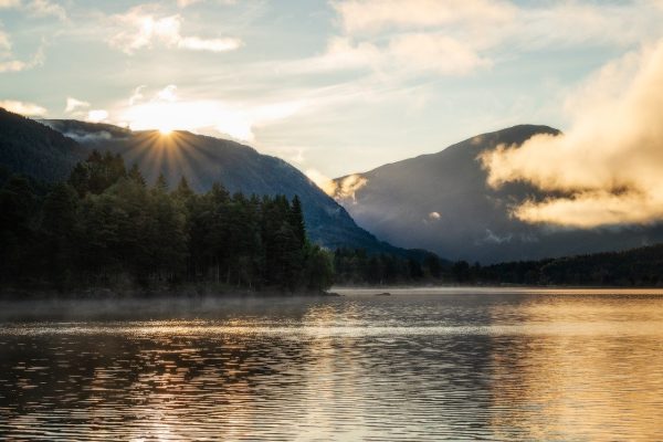 Sunrise over a serene lake surrounded by forested mountains and soft clouds.