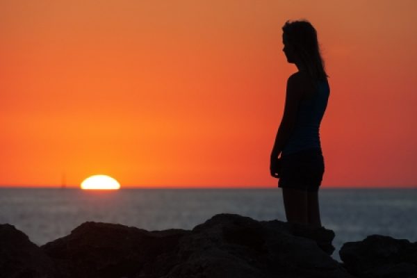 Silhouette of a person standing on rocks, watching the sunset over the ocean with a sailboat in the distance.