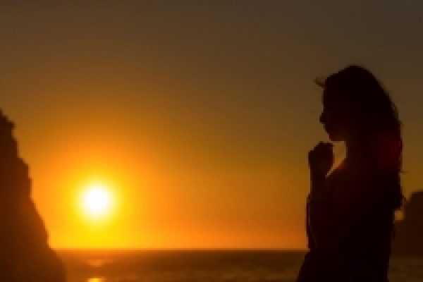 Silhouette of a person looking at the sunset over the ocean near rocky cliffs.
