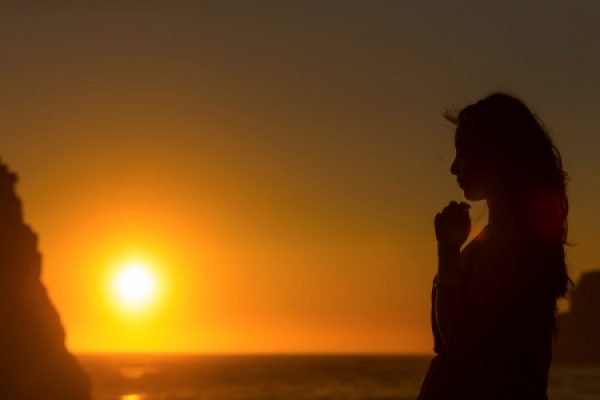 Silhouette of a person by the ocean at sunset, with hands clasped near their face.