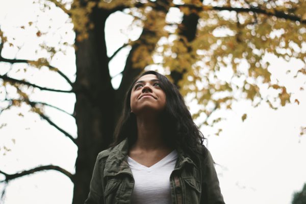 young woman with brown skin and long black hair standing in a forest looking up and slightly smiling