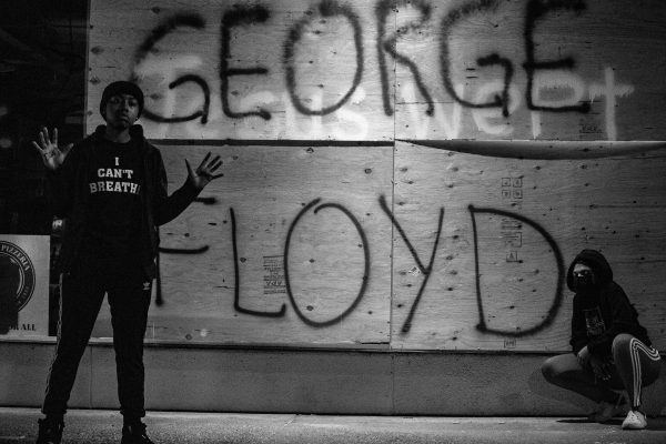 Two people by a wall with George Floyd spray-painted; one raises hands, wearing I cant breathe hoodie.