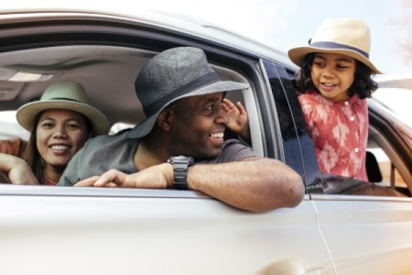 Smiling family wearing hats in a car, with a child leaning out of the window on a sunny day.