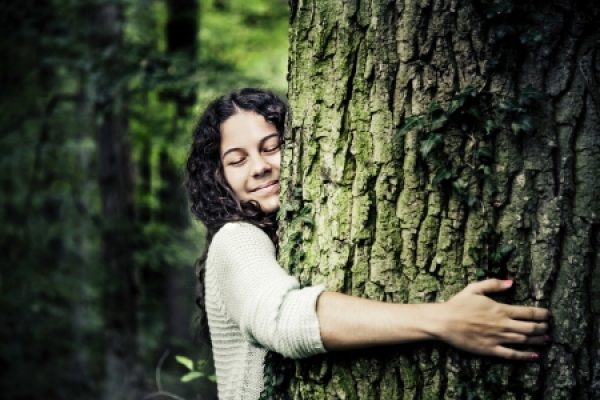 A person with curly hair hugs a tree in a lush green forest, smiling with eyes closed.