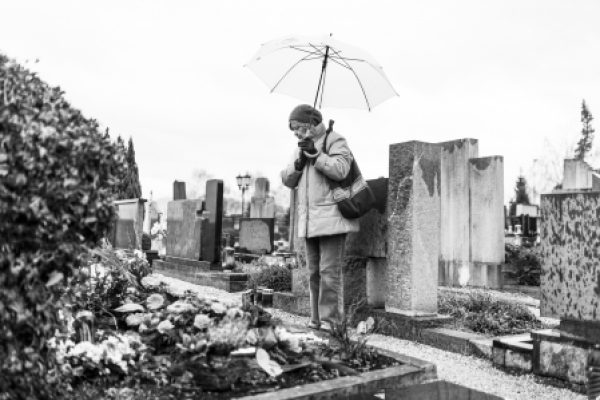 An elderly woman with an umbrella stands thoughtfully by a grave in a cemetery.