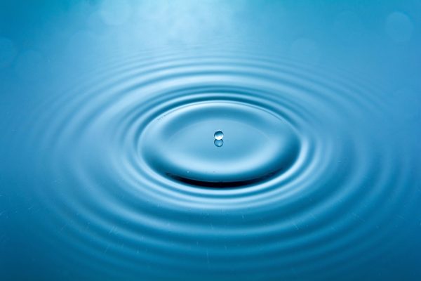 A water droplet falls into a calm blue surface, creating circular ripples.