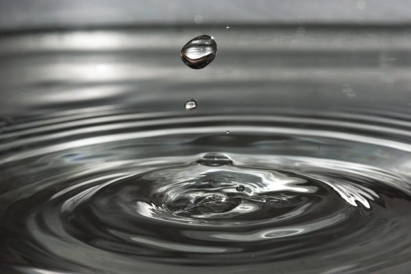 Water droplet splashing on a reflective surface, creating concentric ripples with another droplet above.