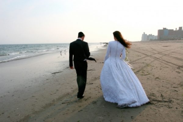 Bride and groom walking on a beach with city buildings in the background.