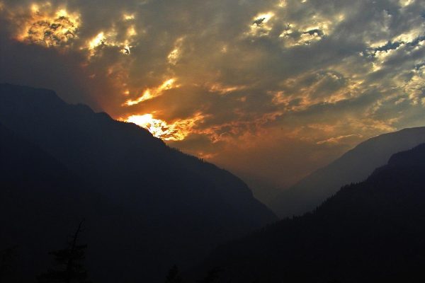 Sunset glows through dramatic clouds over a dark mountain valley, creating a striking silhouette.