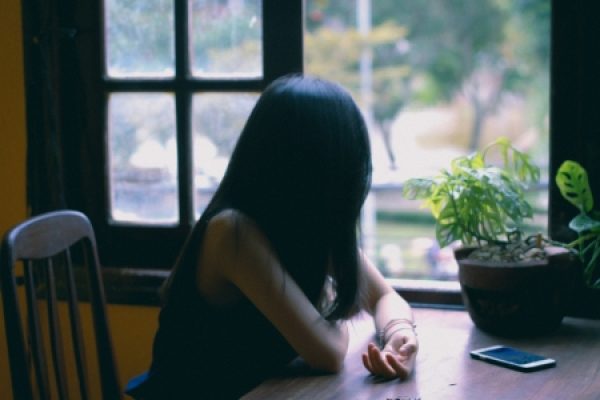 Person sitting at a table by a window, looking outside, with a smartphone and a potted plant nearby.