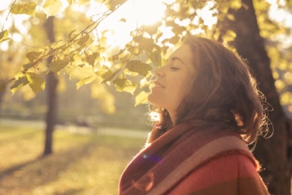 Woman with eyes closed enjoying sunlight, standing near tree with autumn leaves, in a park.
