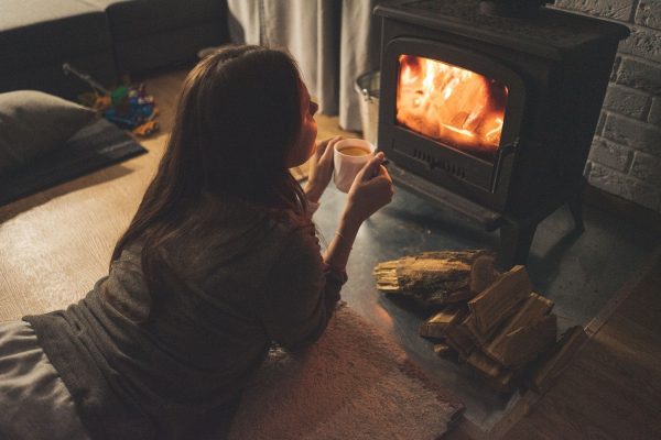 A person lying on a rug, holding a mug, and watching a wood-burning stove with logs nearby.