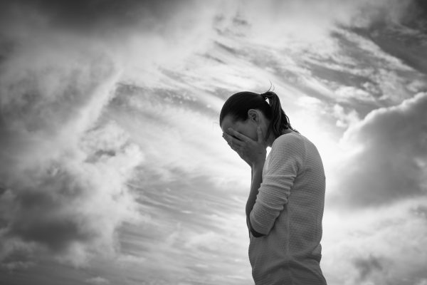 Silhouetted woman covers her face with hands against a dramatic, cloudy sky in black and white.