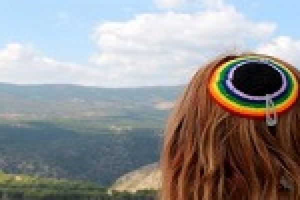 Person with rainbow kippah looking at a scenic mountain view under a partly cloudy sky.