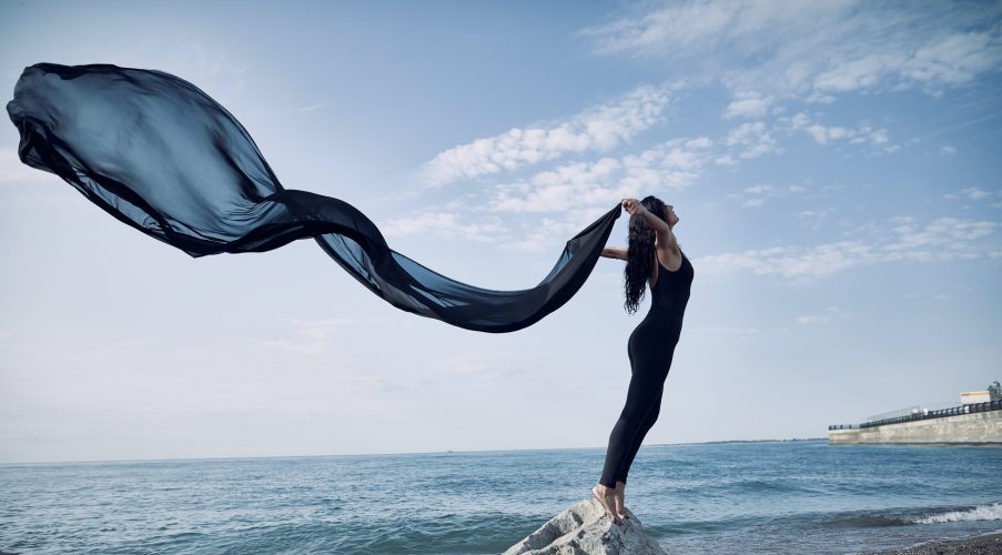 woman standing on a rock by the ocean with black scarf flowing behind her sm