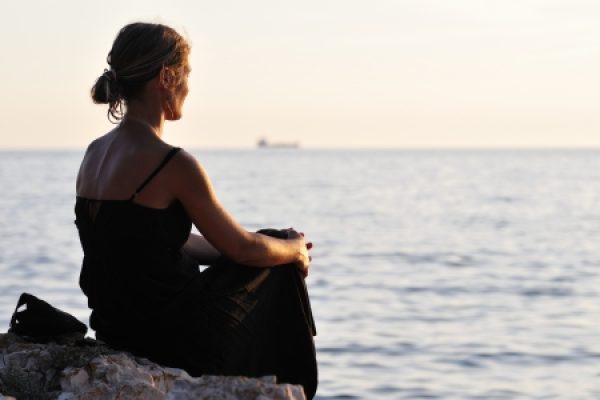 A woman in a black dress sits on a rock, gazing at the ocean during sunset.