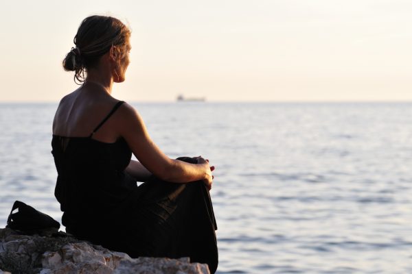 Woman in a black dress sitting on a rock, gazing at the sea during sunset.