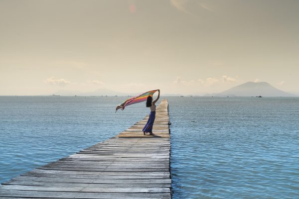 Person holding a colorful scarf stands on a wooden pier over calm blue water with mountains in the distance.