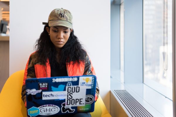 Person in camouflage cap and orange jacket using a laptop covered in stickers, seated by a window.