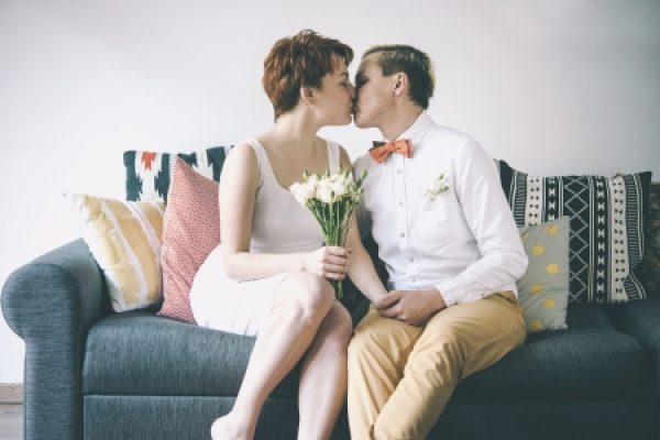 A couple sitting on a sofa, kissing while holding a bouquet of white flowers, with patterned pillows in the background.