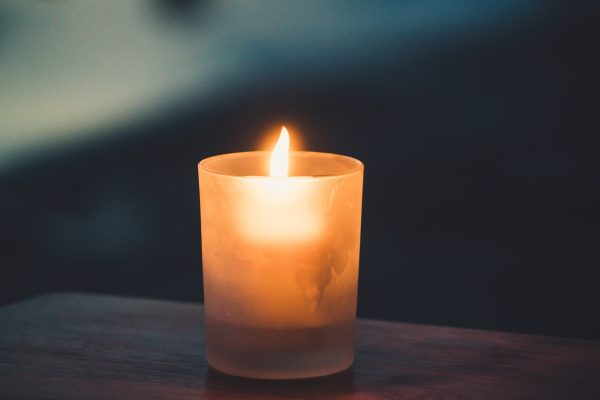 A lit candle in a frosted glass holder casts a warm glow in a dark setting.