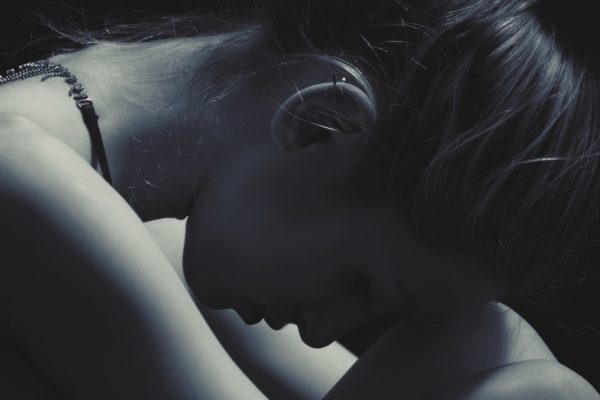 Silhouette of a woman with her head down, sitting and embracing her knees in a dimly lit setting.